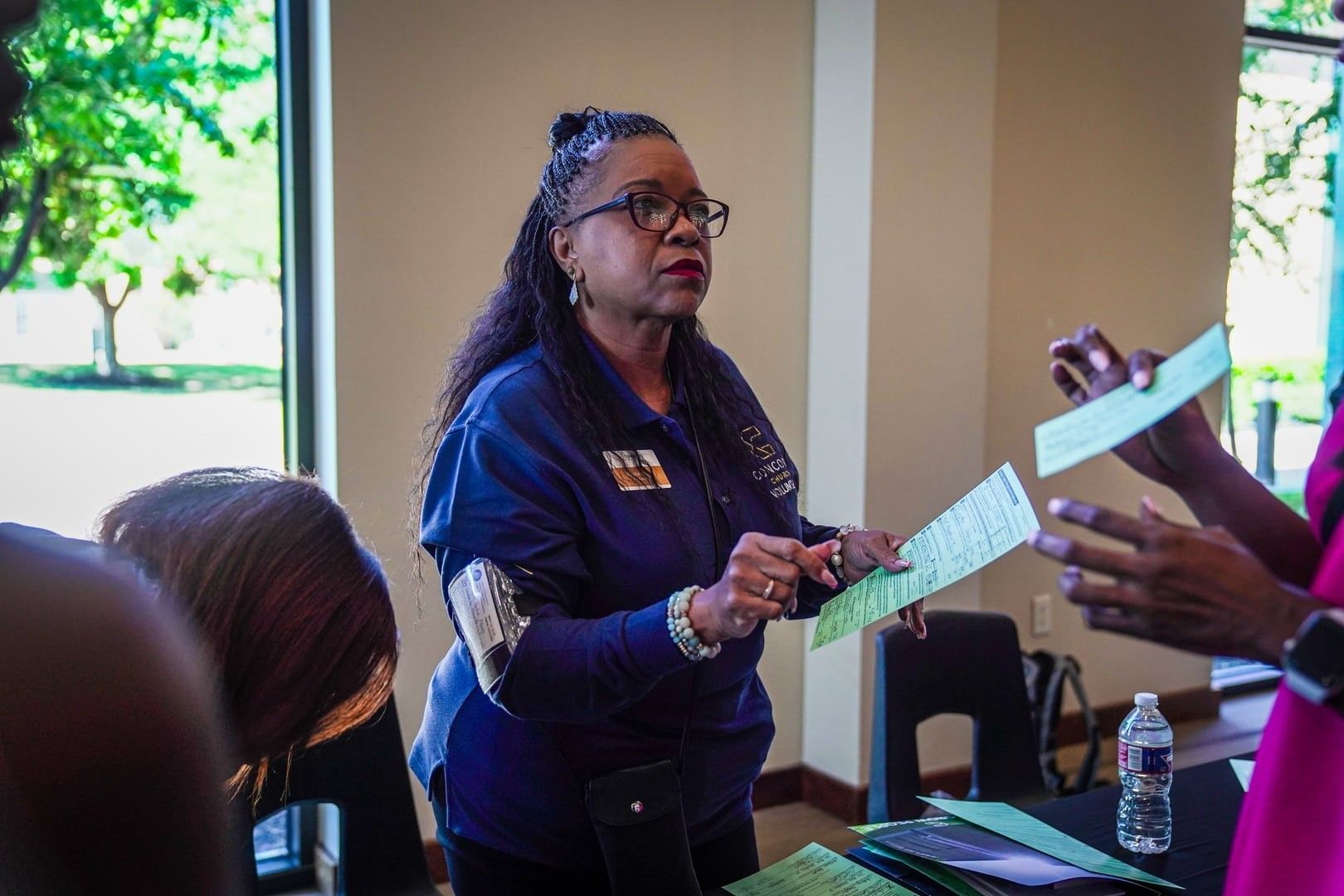 Woman in blue shirt, handing out green papers in a room. Others nearby.