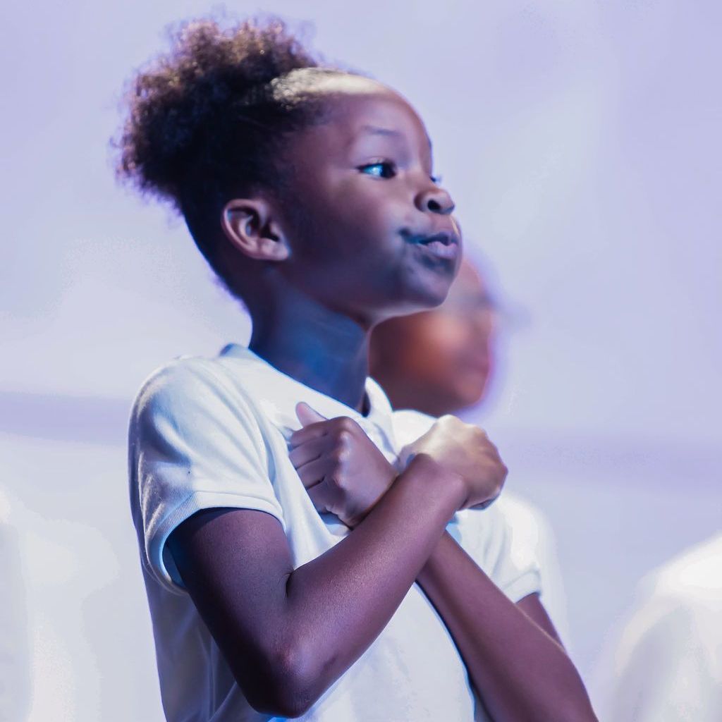 Young Black girl singing on stage with hands crossed over chest.
