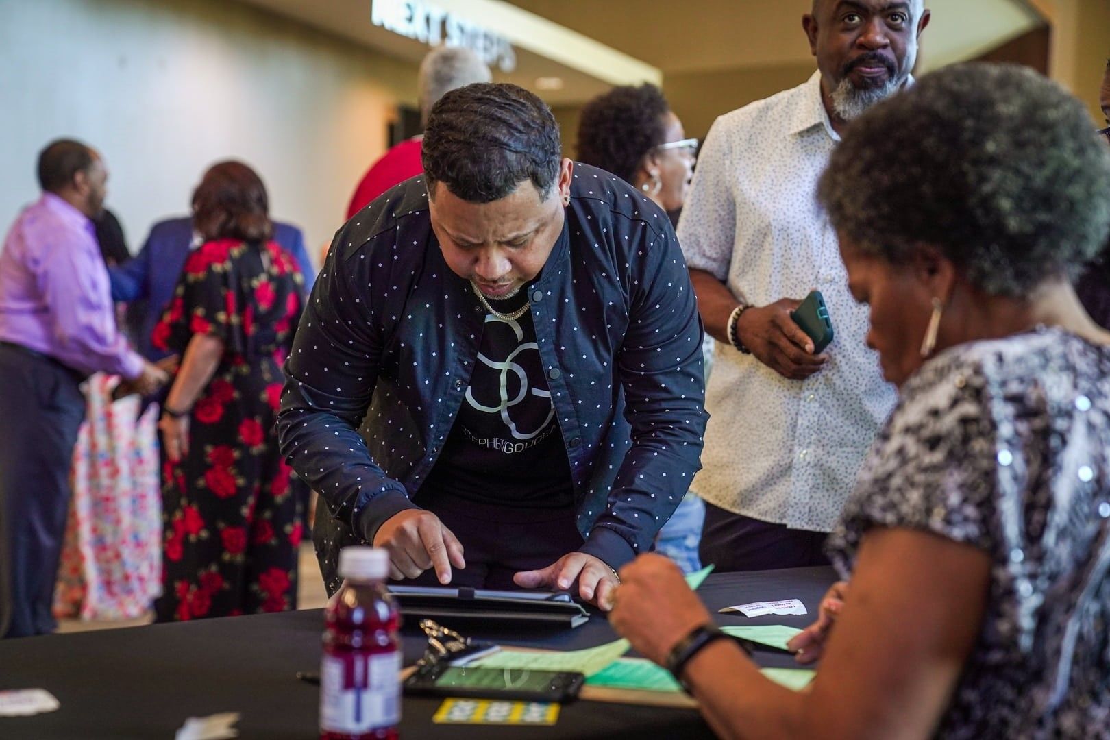 People at a registration table, one man using a tablet, another woman writing; event setting.