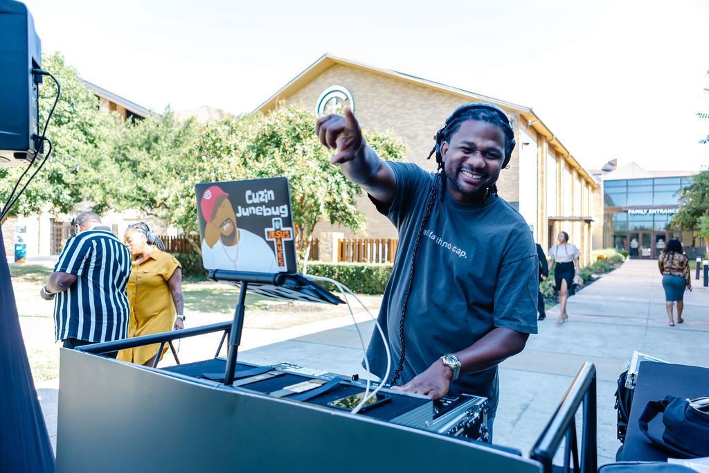 A smiling DJ raises a hand holding a logo. In a courtyard, he stands behind a DJ setup.