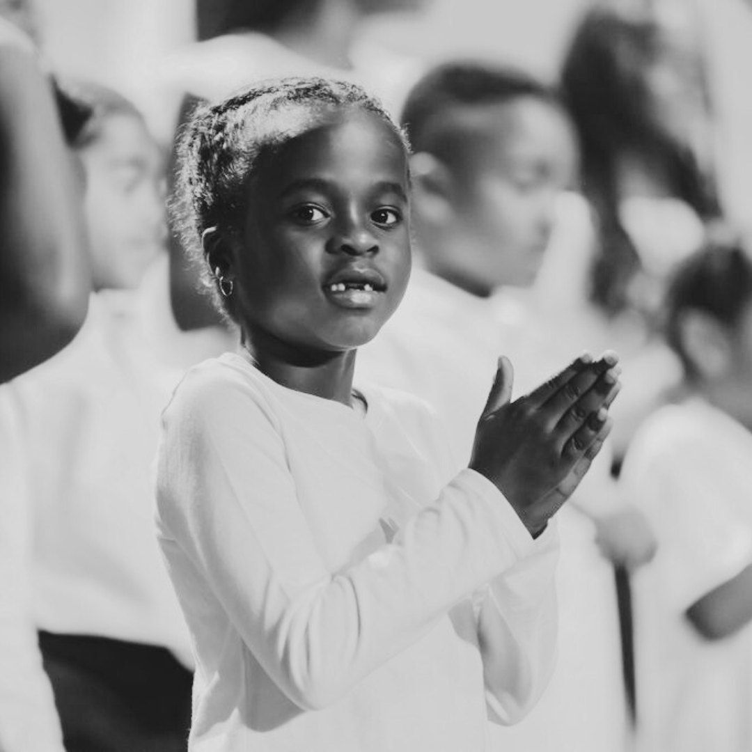 Young girl clapping, smiling, in a group of people; wearing white clothing.