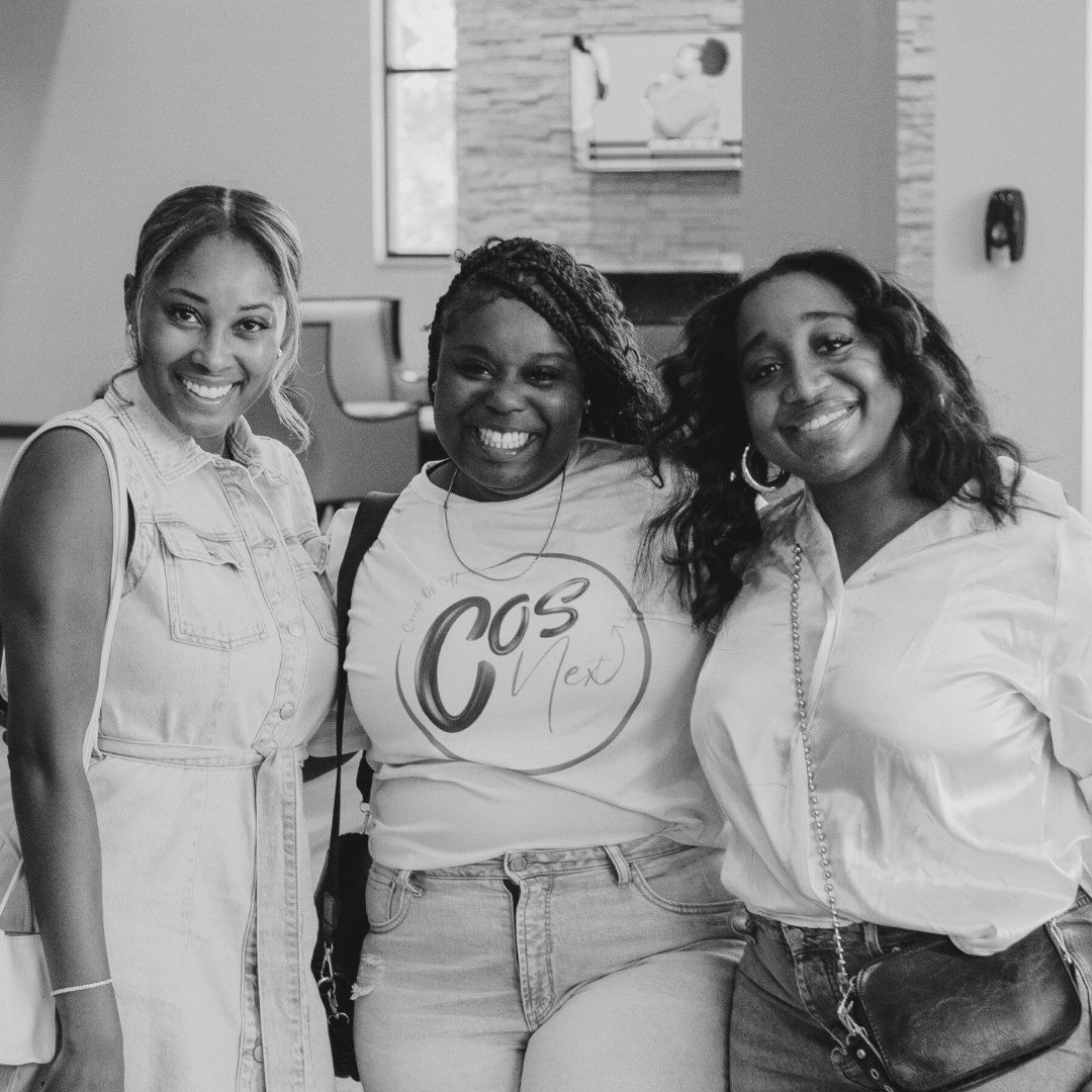 Three smiling women posing together indoors; one in a denim dress, one in a t-shirt, and one in a button-down shirt.