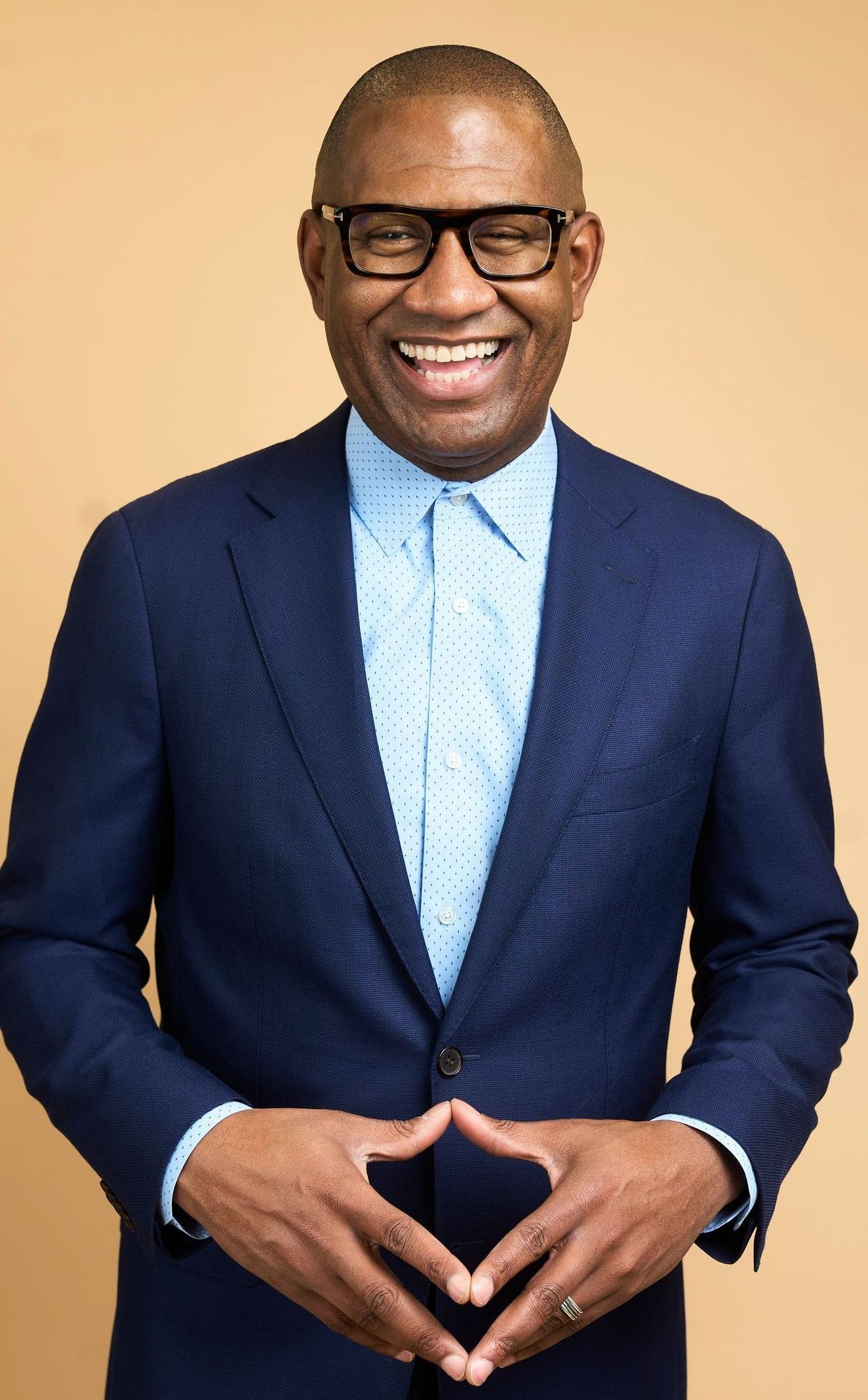 Smiling Black man in navy blazer, light blue patterned shirt, hands clasped, against tan backdrop.