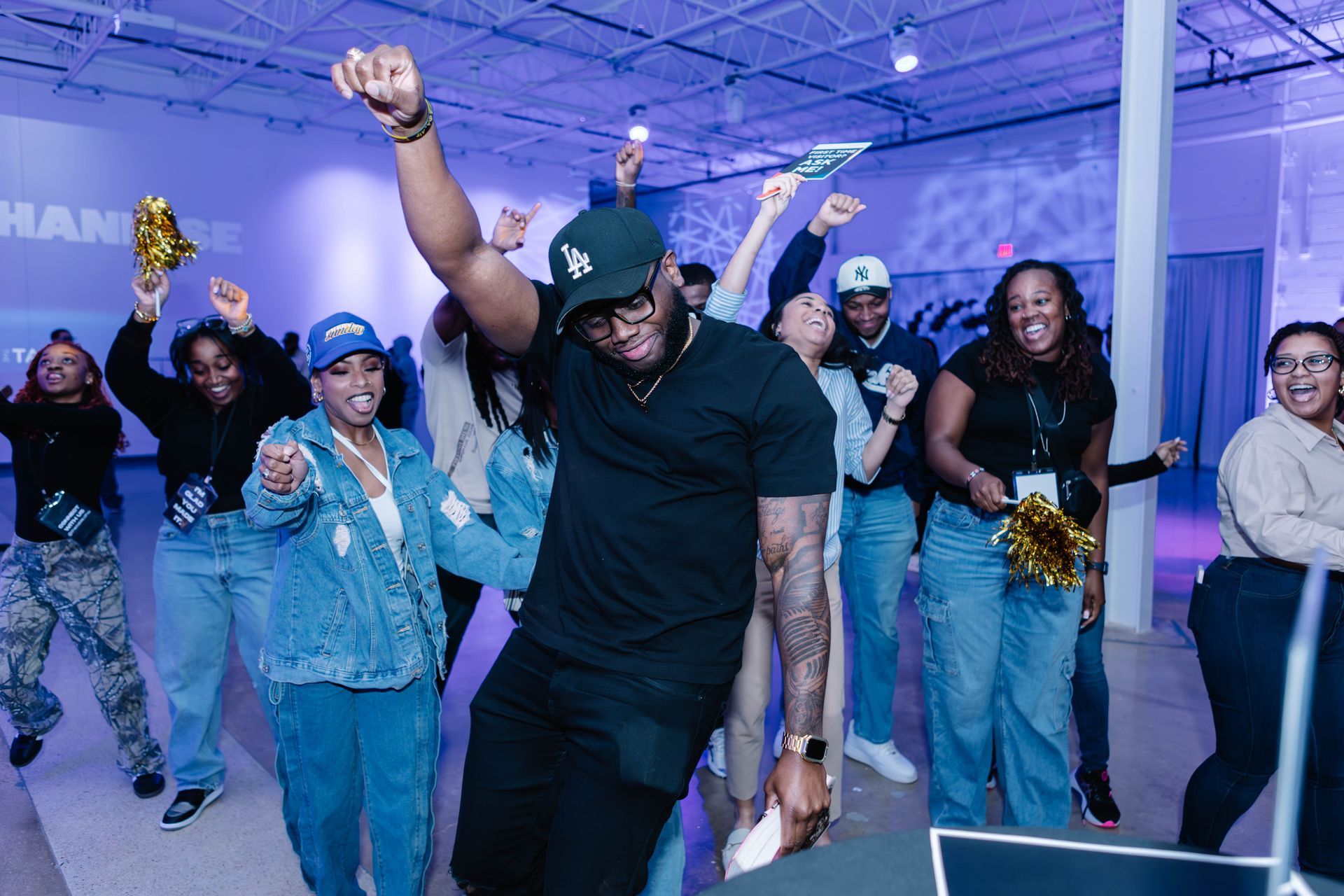 Group of people dancing joyfully in a brightly lit space. Some hold gold pom-poms, smiling.