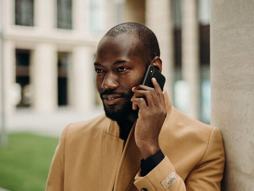 A man in a tan coat is leaning against a wall while talking on a cell phone.