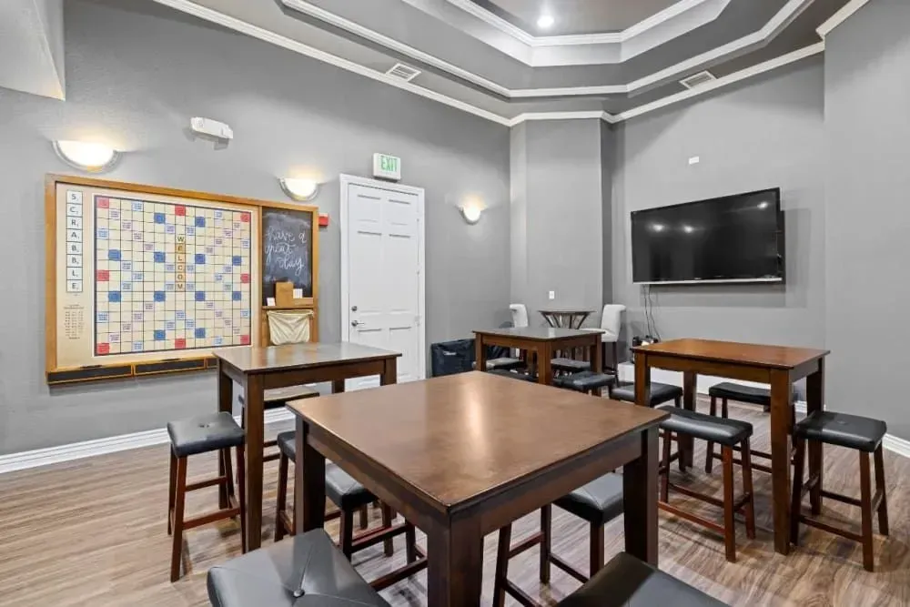 A community room with gray walls, wood tables, black stools, a TV, and a mounted Scrabble board.