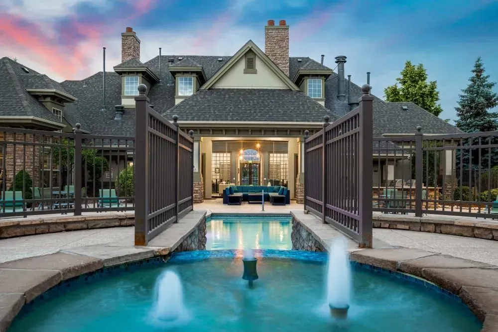 Outdoor pool with fountains in front of a luxury home with a dark roof and brick accents.