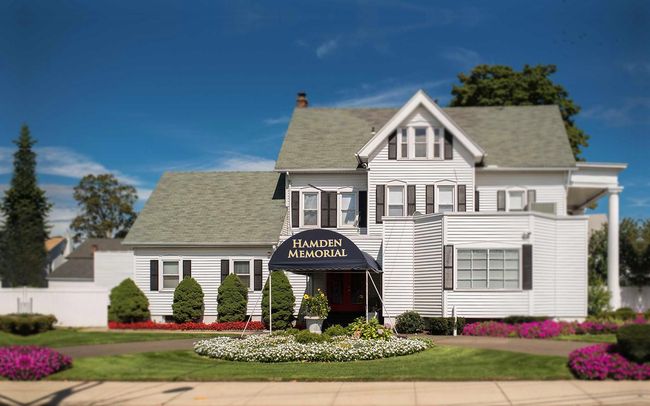 White Victorian-style house with black awning, flower beds, and manicured lawn under a blue sky