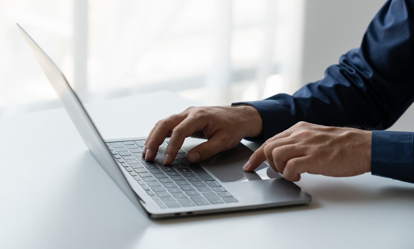 Hands typing on a silver laptop at a bright desk near a window.