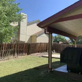Backyard with wooden fence, covered patio, grass, and a beige house under a blue sky.