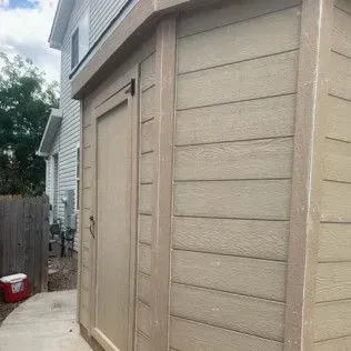Tan shed with wood-like siding and trim, next to a house with gray siding.