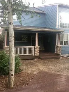 Blue house with brown porch, white railing, and stone columns. Tree in foreground.