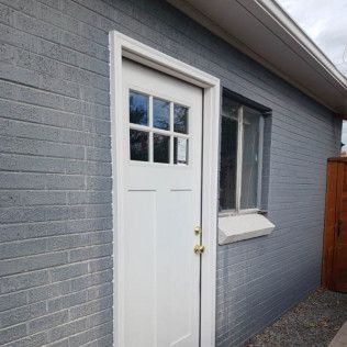White door with glass panes and window next to it, set in a gray brick building, with white trim.