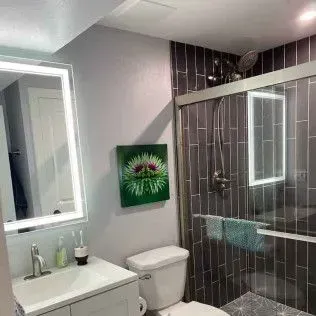 Bathroom with gray walls and tile, a white vanity, toilet, and a glass-enclosed shower with a painting.