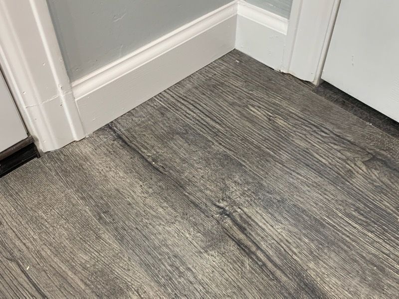 Corner of a room with gray wood-look flooring and white trim, featuring a door frame and wall.