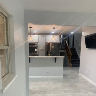 A renovated kitchen and living area, featuring light gray cabinets, a breakfast bar, and stairs in the background.