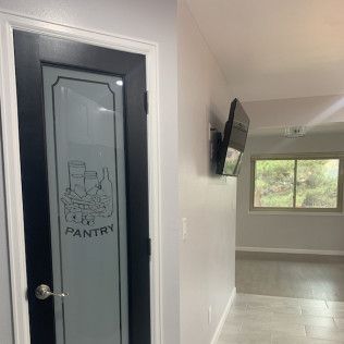 Pantry door with frosted glass and pantry label, black frame. Room with TV mounted on wall and window.