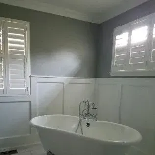 Bathroom with white wainscoting, a clawfoot tub, and shutters on the windows; gray walls.