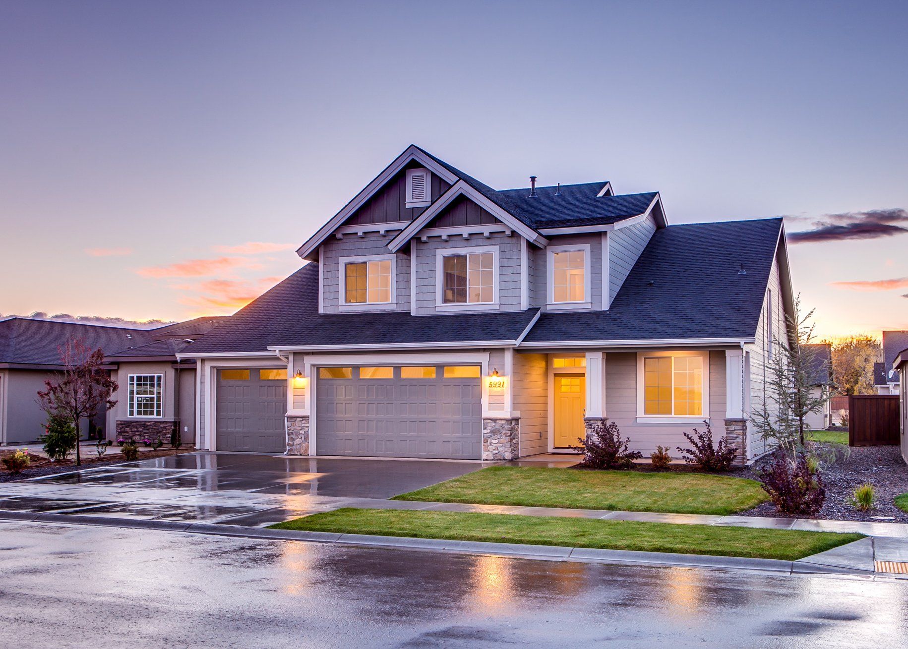 Two-story house with gray siding, two-car garage, and dark roof at dusk with wet street.