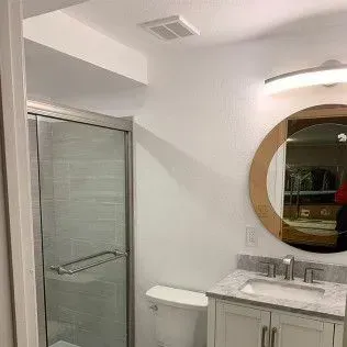 Bathroom with a glass shower, toilet, vanity, round mirror, and recessed lighting. White walls and cabinets.