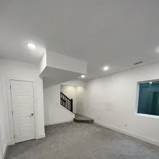 Empty, carpeted room with a staircase leading up and a door on the left; white walls and ceiling.