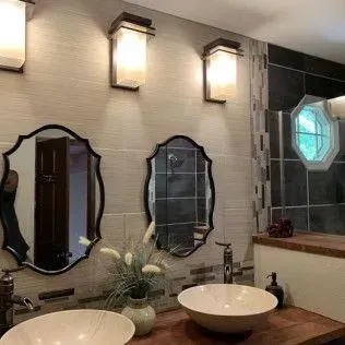 Bathroom with two sinks, mirrors, lights, and a shower with a window. Brown and beige tones.