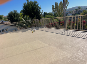 Sunlit concrete rooftop patio with metal railing and potted plants, overlooking trees and hills