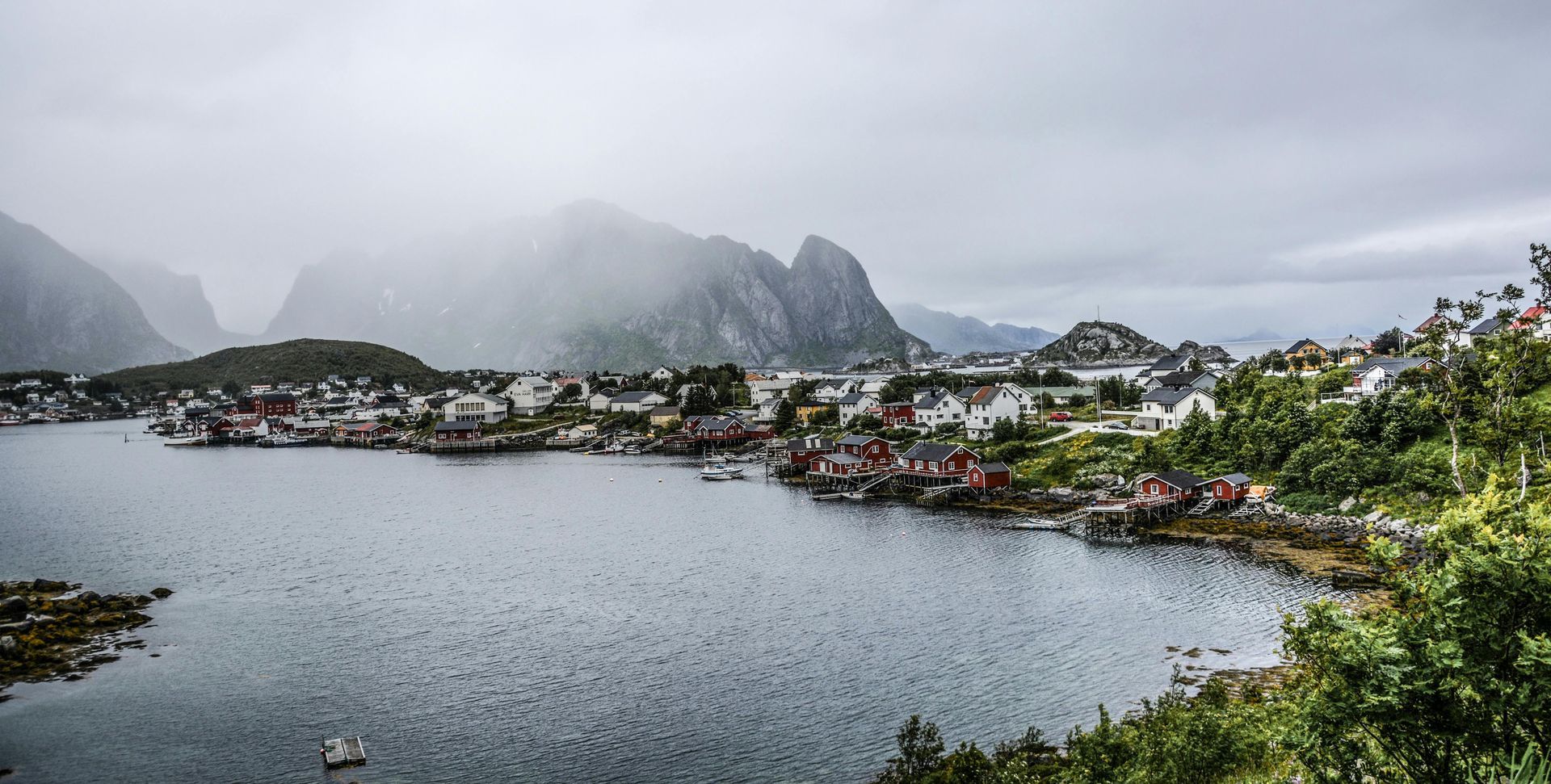 Townscape on a fjord under a cloudy sky, mountains in the background. Buildings, water, and green foliage.