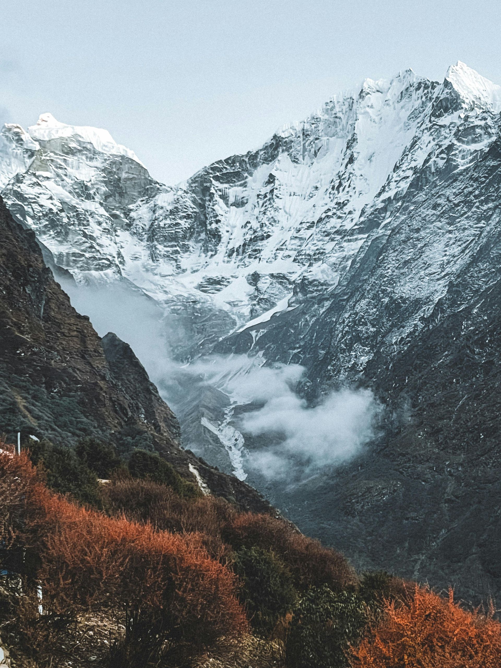 Snow-covered mountain peaks with a valley in between, cloudy sky, autumn trees.