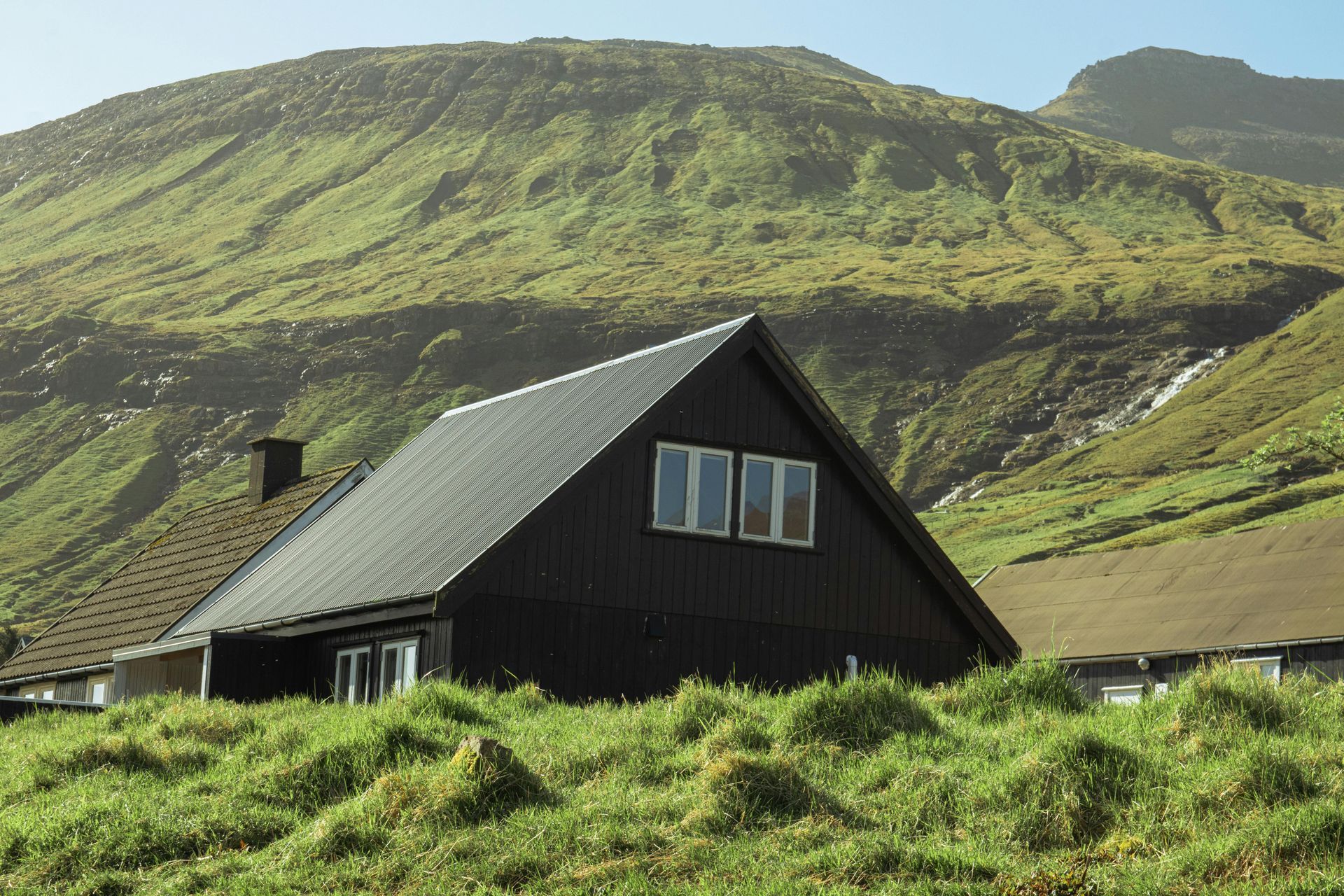 A dark cabin with a gray roof sits in grassy terrain, backed by a large green hillside.