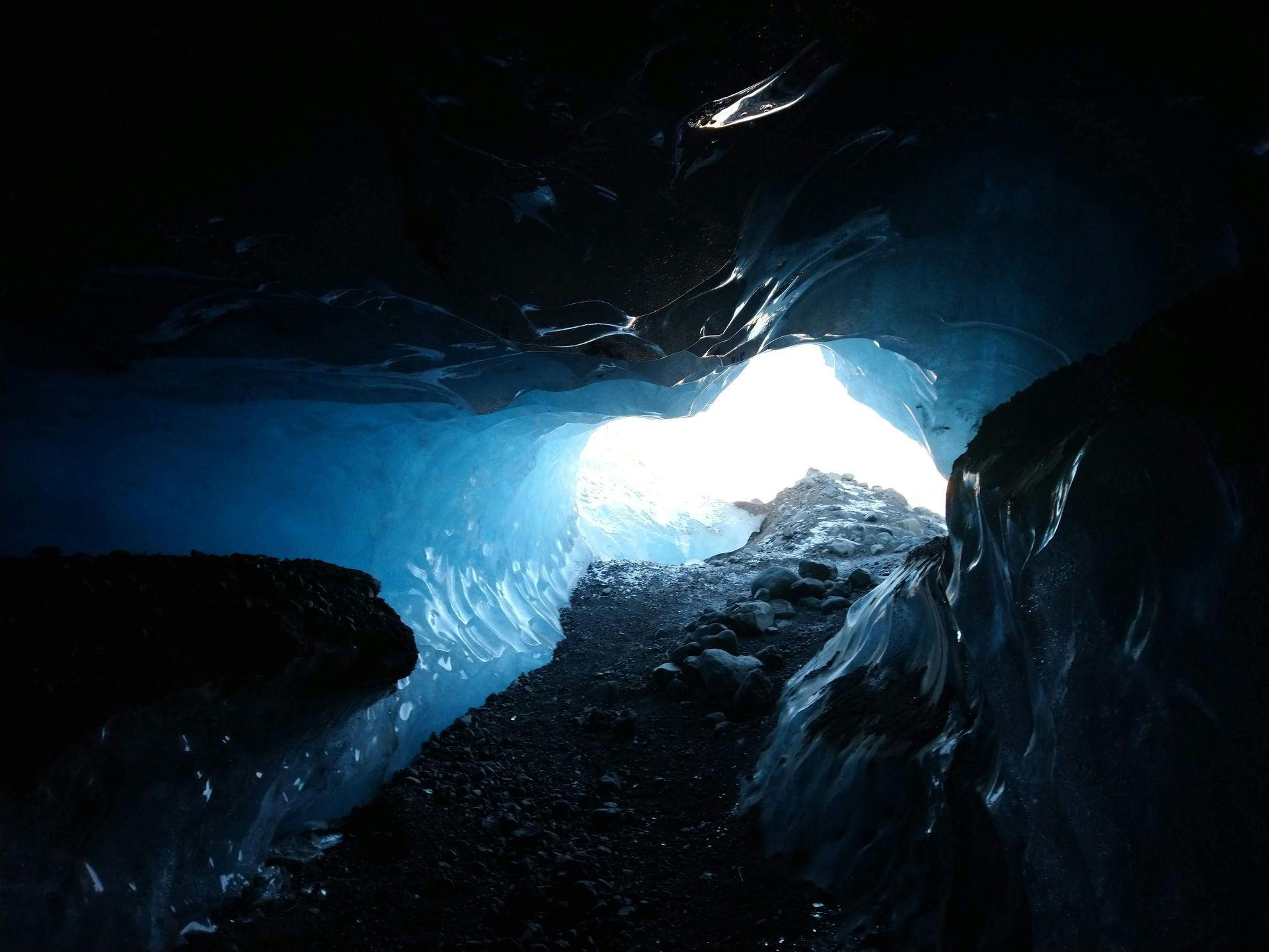 Inside a blue ice cave, looking towards the bright opening. Dark passage with icy walls.