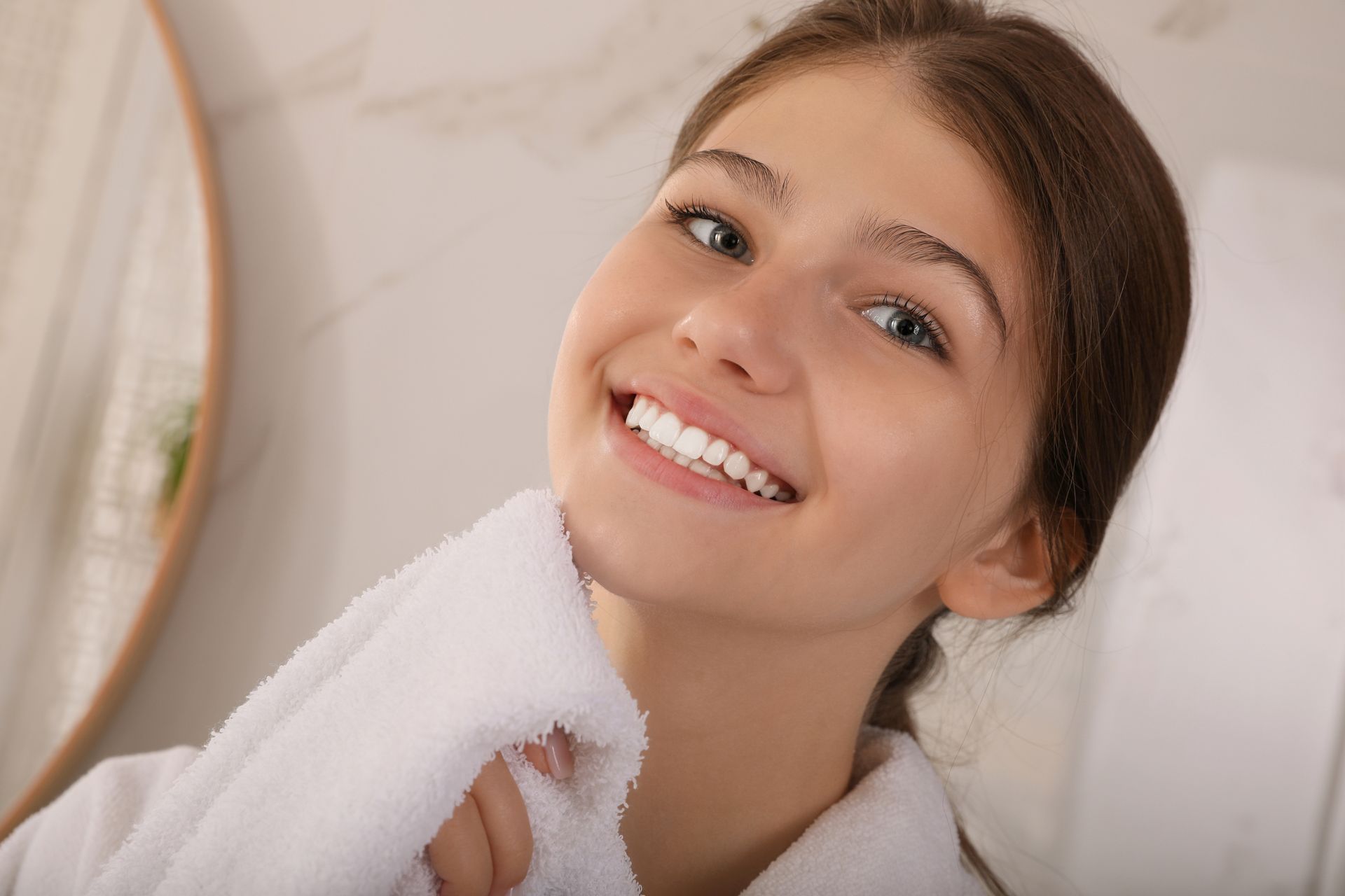 A young woman with clear skin and a bright smile holds a white towel to her face. 