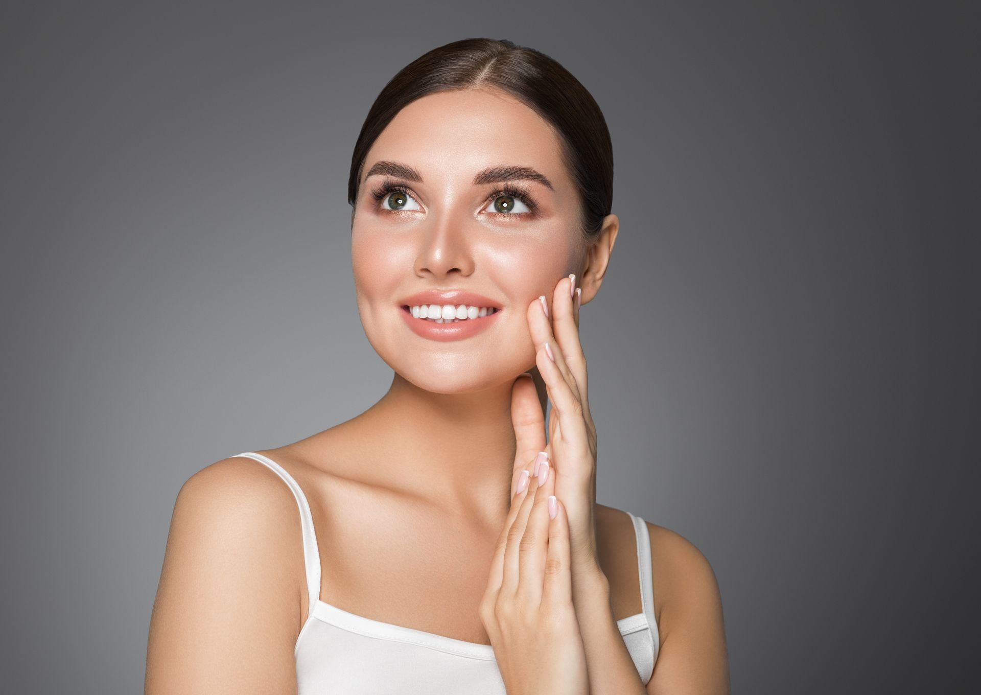 Smiling woman in a white top touching her cheek against a gray background