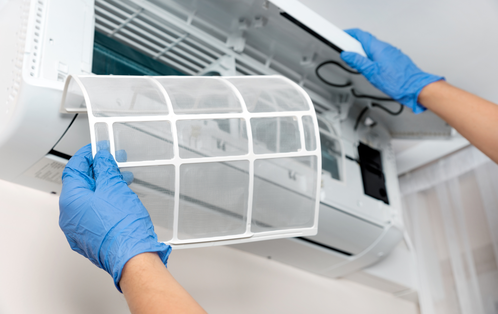 Hands in blue gloves removing an air conditioner filter. White unit on a wall.