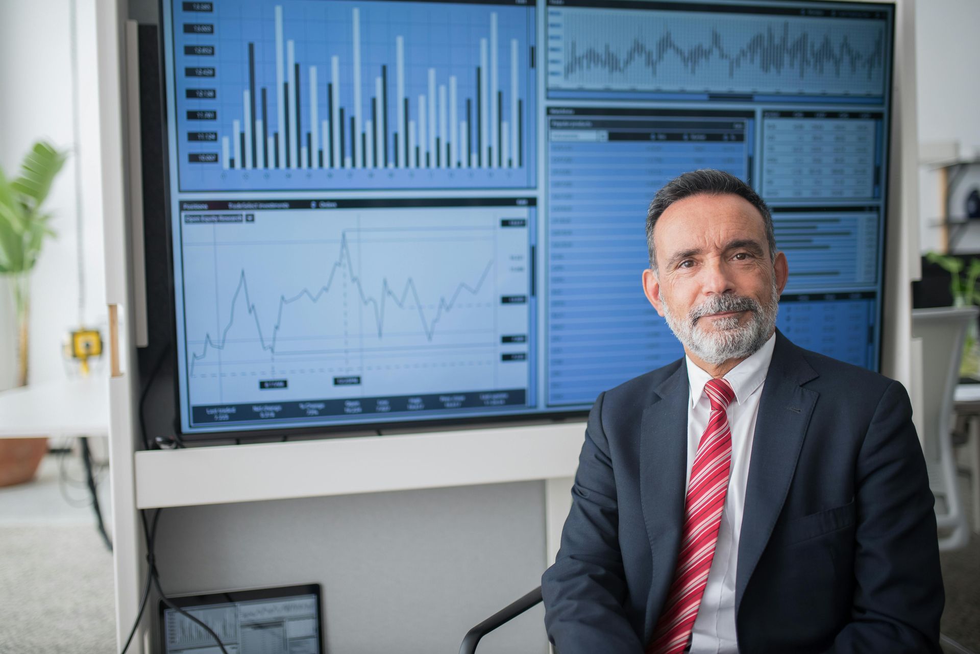 Businessman in a suit seated beside screens showing financial charts and graphs