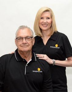 Two people in black polos posing against a white background, one seated and one standing with a hand on his shoulder