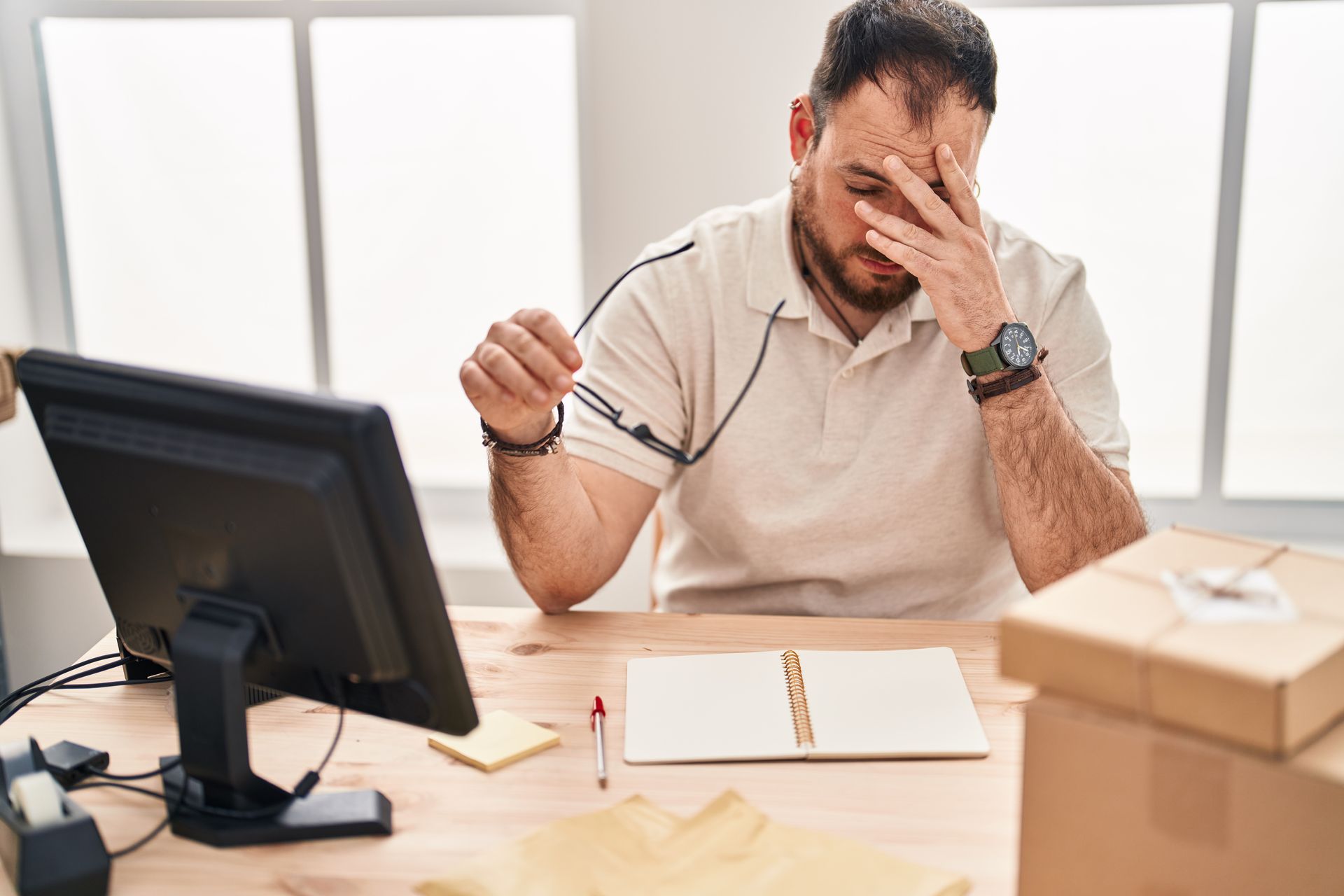 Stressed man at desk with hand on face, holding glasses near a computer and moving boxes