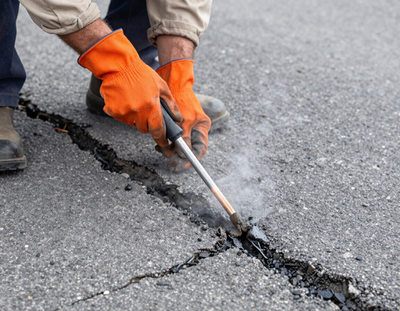 Worker using a pry bar to repair a cracked asphalt road, wearing orange gloves.