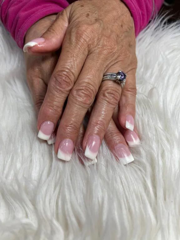 Manicured hands with a French tip style, resting on a white faux fur surface, with a ring on the middle finger.