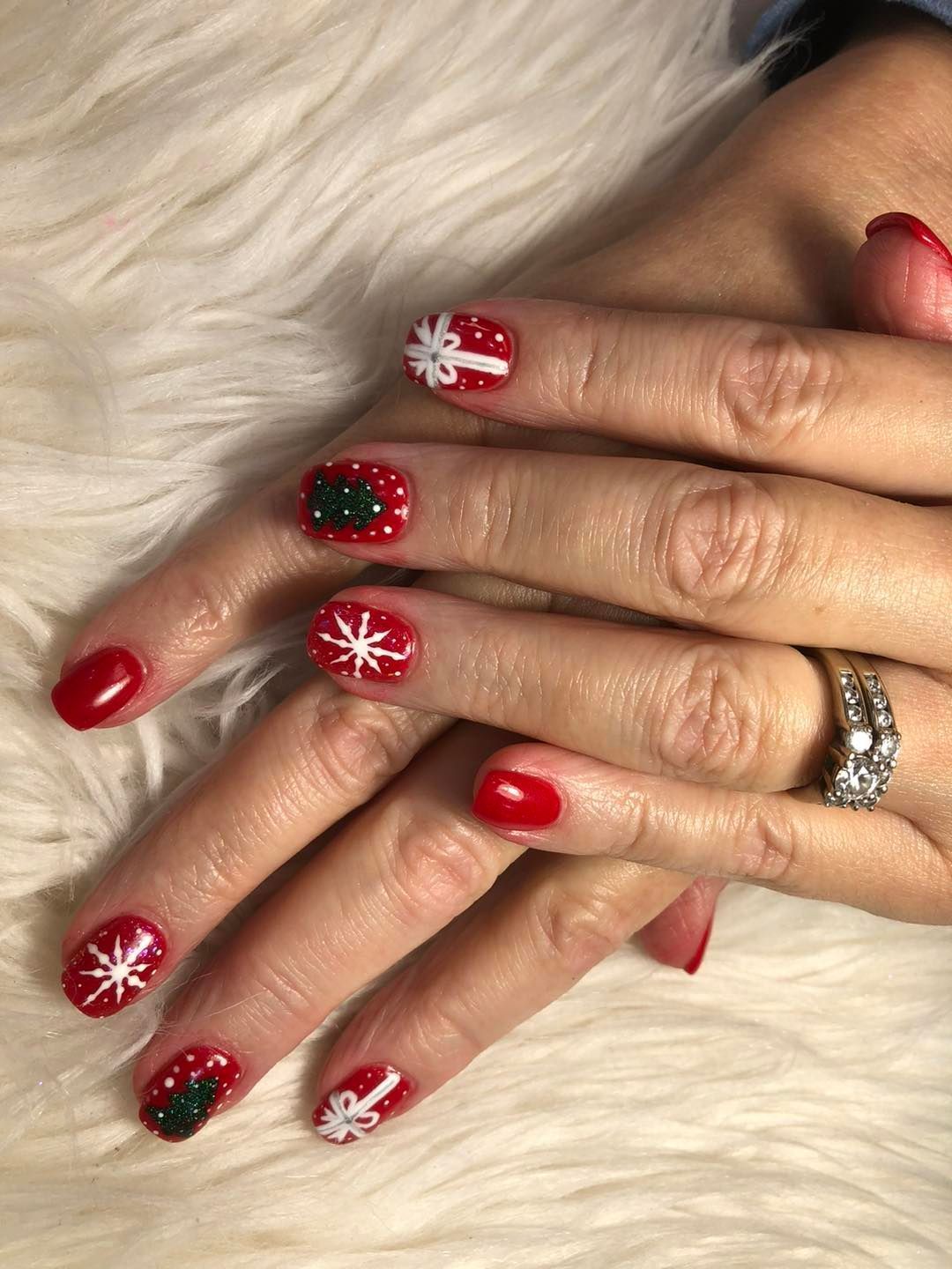 Manicured fingernails with red polish and festive white snowflakes, bows, and green Christmas tree nail art.
