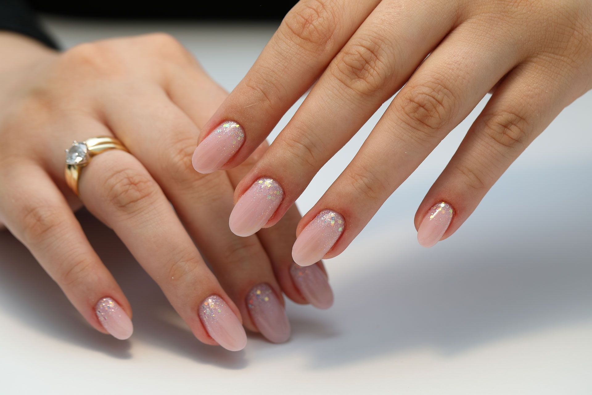 Close-up of hands with neatly manicured, pale pink nails with subtle glitter tips and a gold ring on the ring finger.