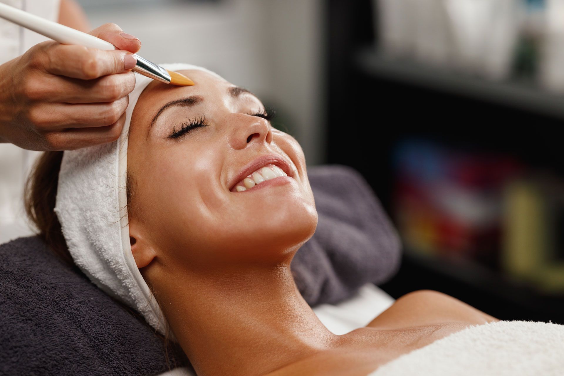 A person with a headband smiles while an esthetician applies a facial treatment with a brush in a spa setting.