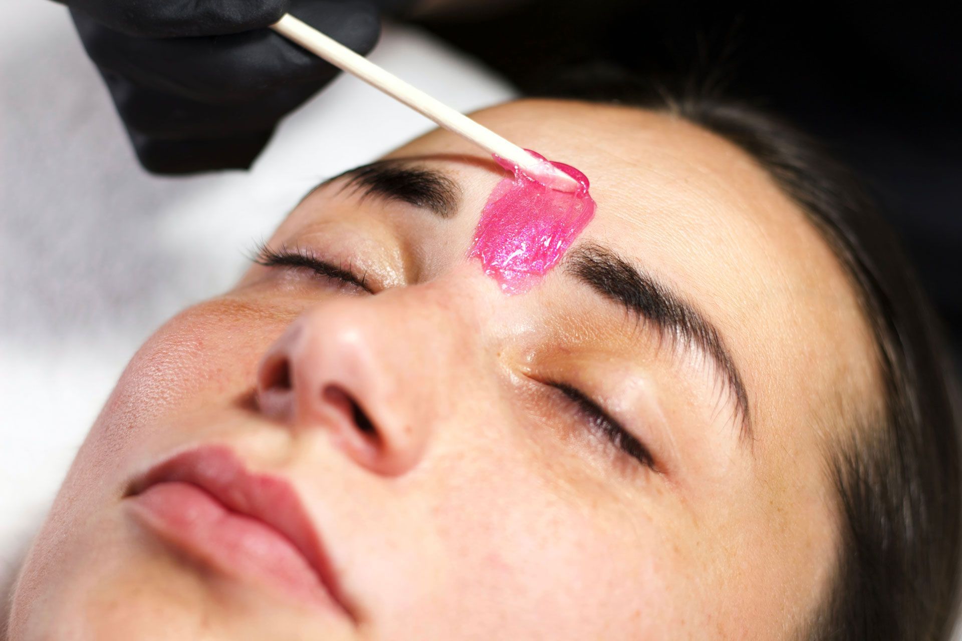 A gloved hand uses a wooden applicator to apply pink wax between the eyebrows of a person lying down.
