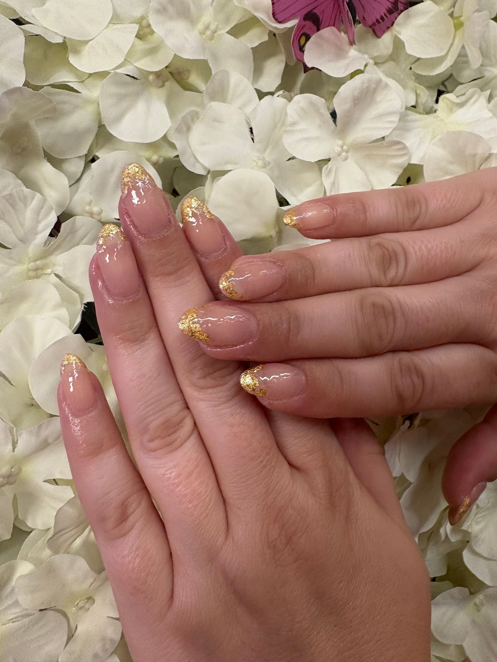 Two hands showing a soft pink manicure with gold foil accents on the tips, set against a background of white flowers.