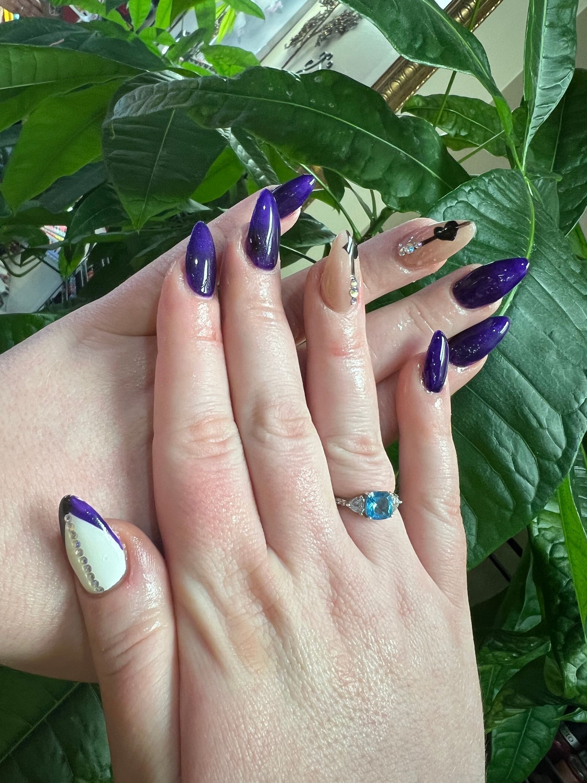 Hands with almond-shaped nails in dark purple and nude, decorated with rhinestones, held against a lush green plant.
