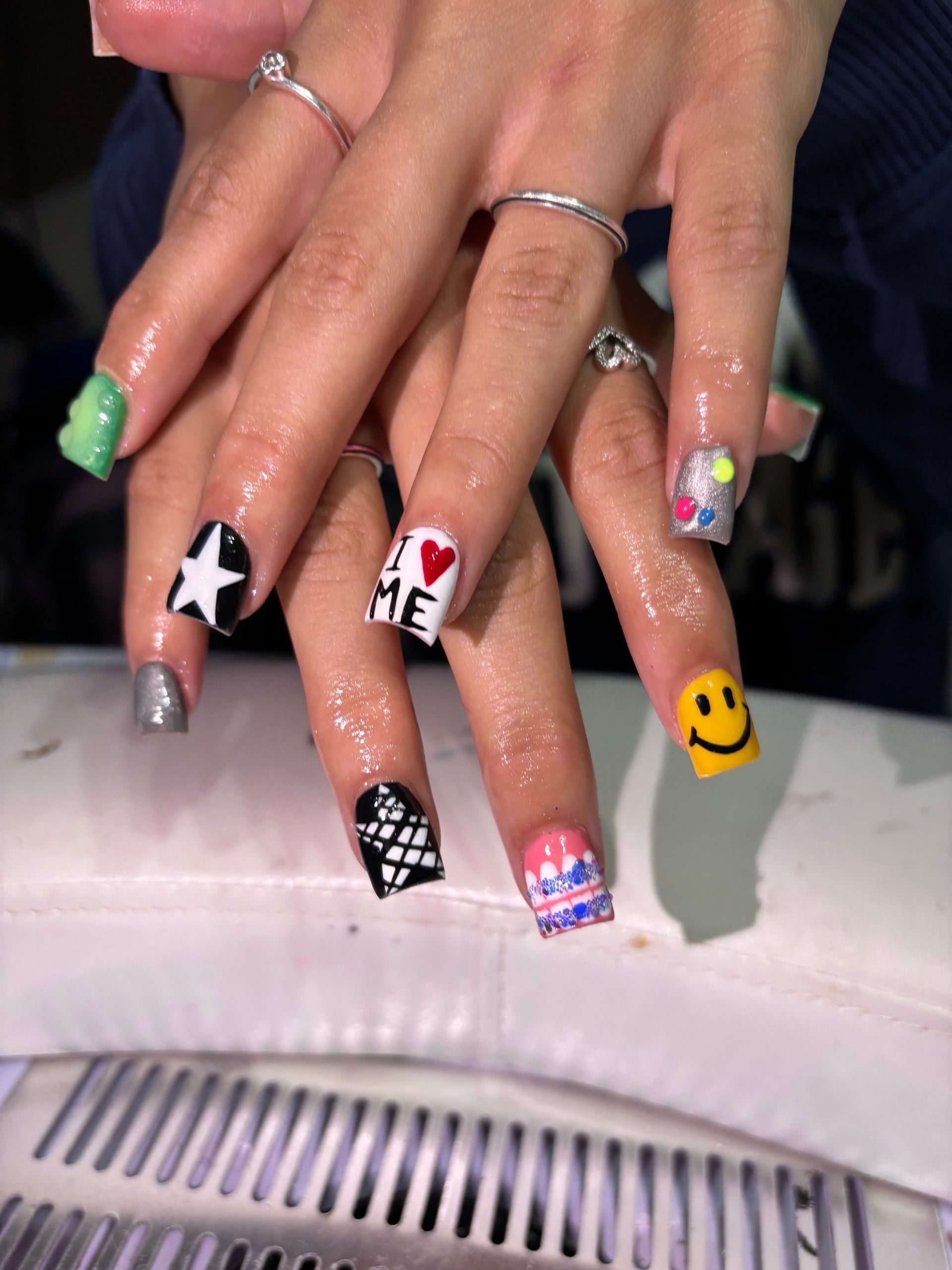 Hands displaying square-shaped nails with colorful, varied designs including a star, a smiley face, and “I heart ME”.