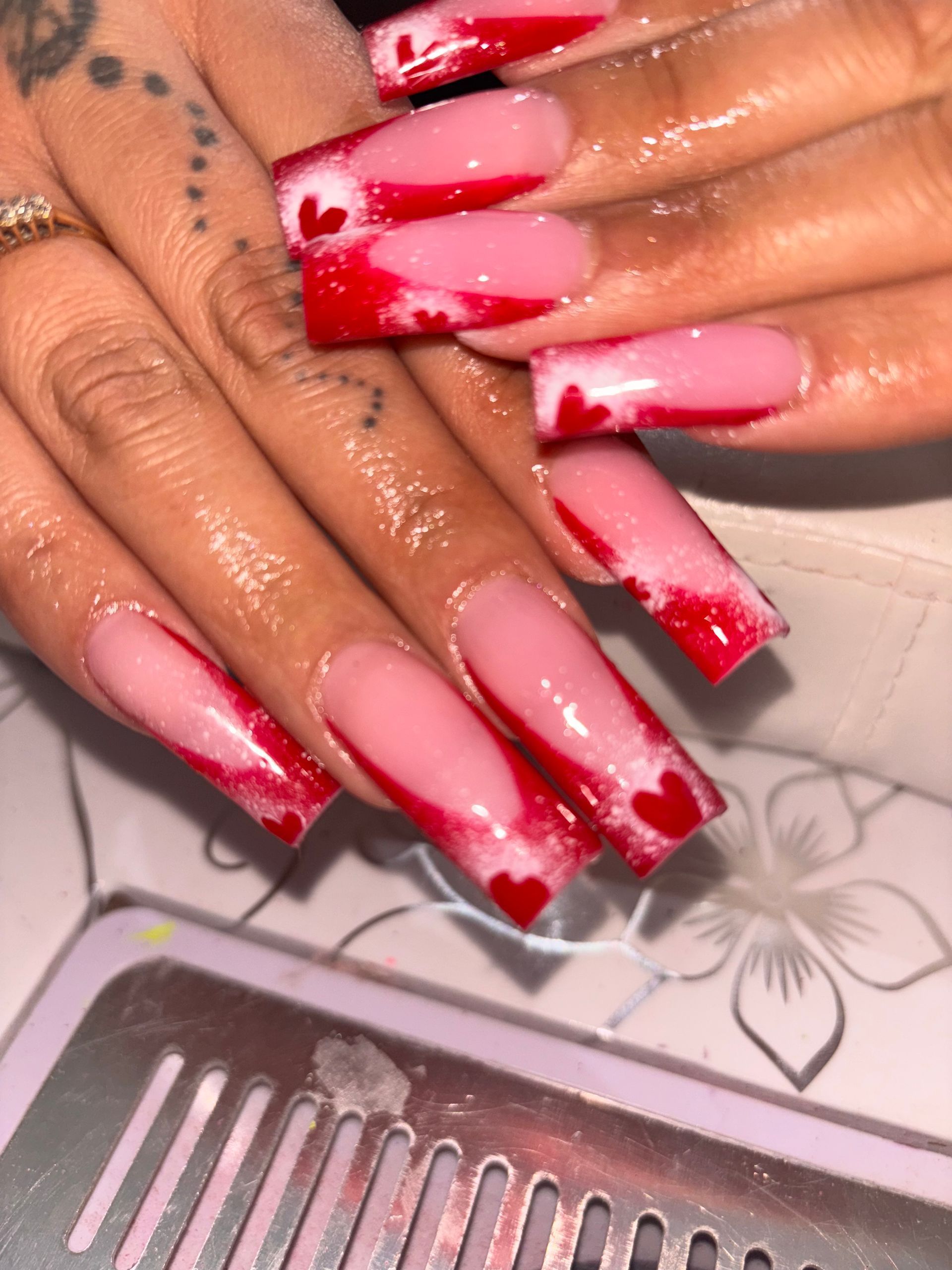 Long square-shaped nails with pink base, red French tips, and small red heart accents on a salon desk.