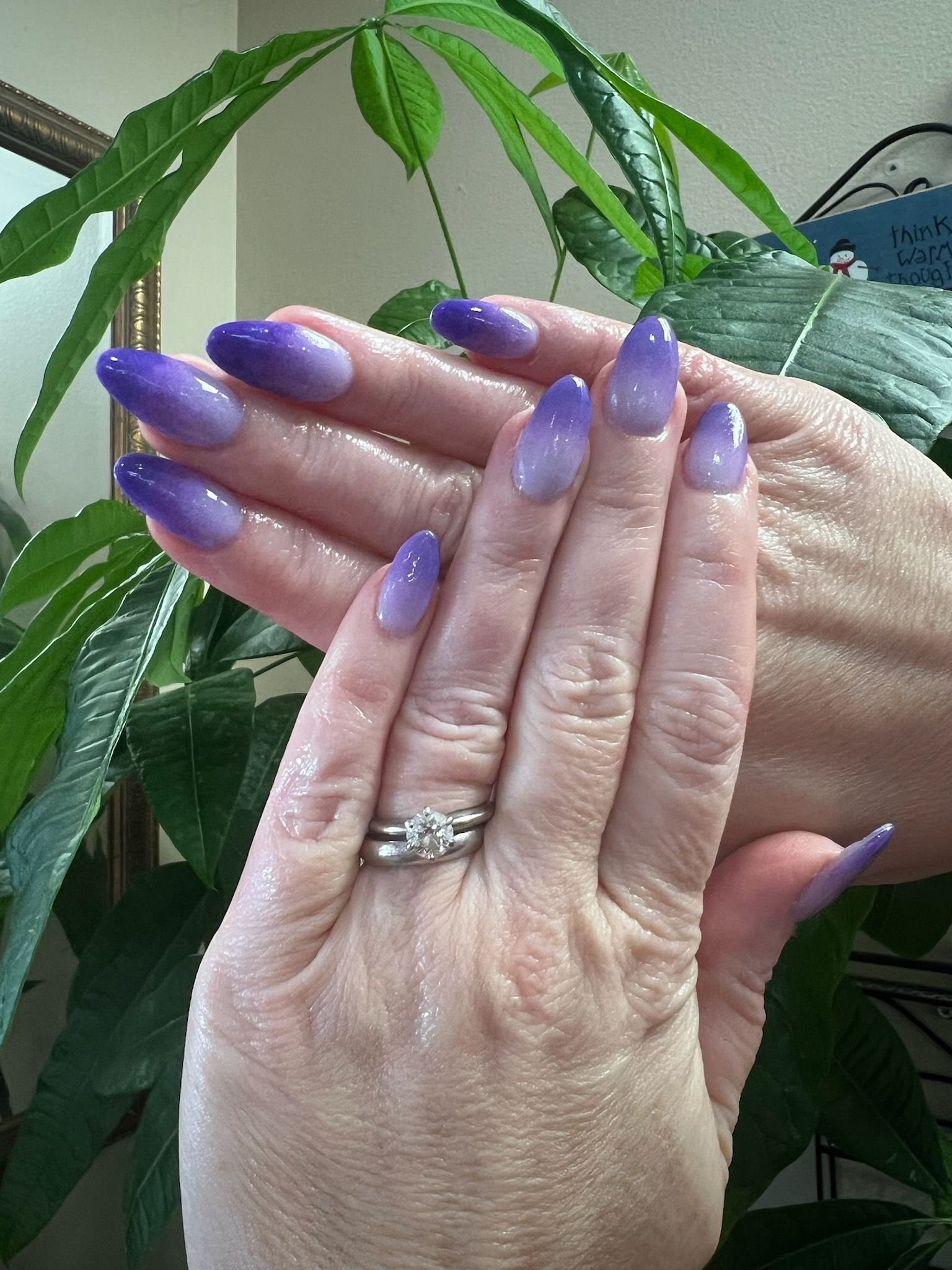 Hands with oval-shaped purple ombre nails, featuring a dark to light gradient, held against a green plant background.