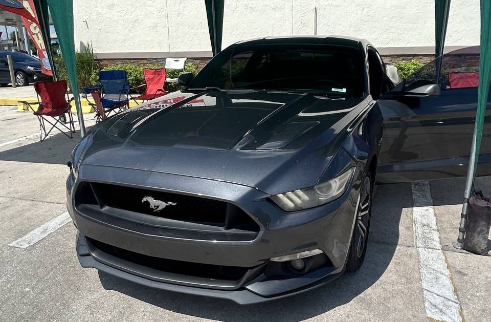 Dark gray Ford Mustang with door open, parked in a parking lot, under a green canopy.