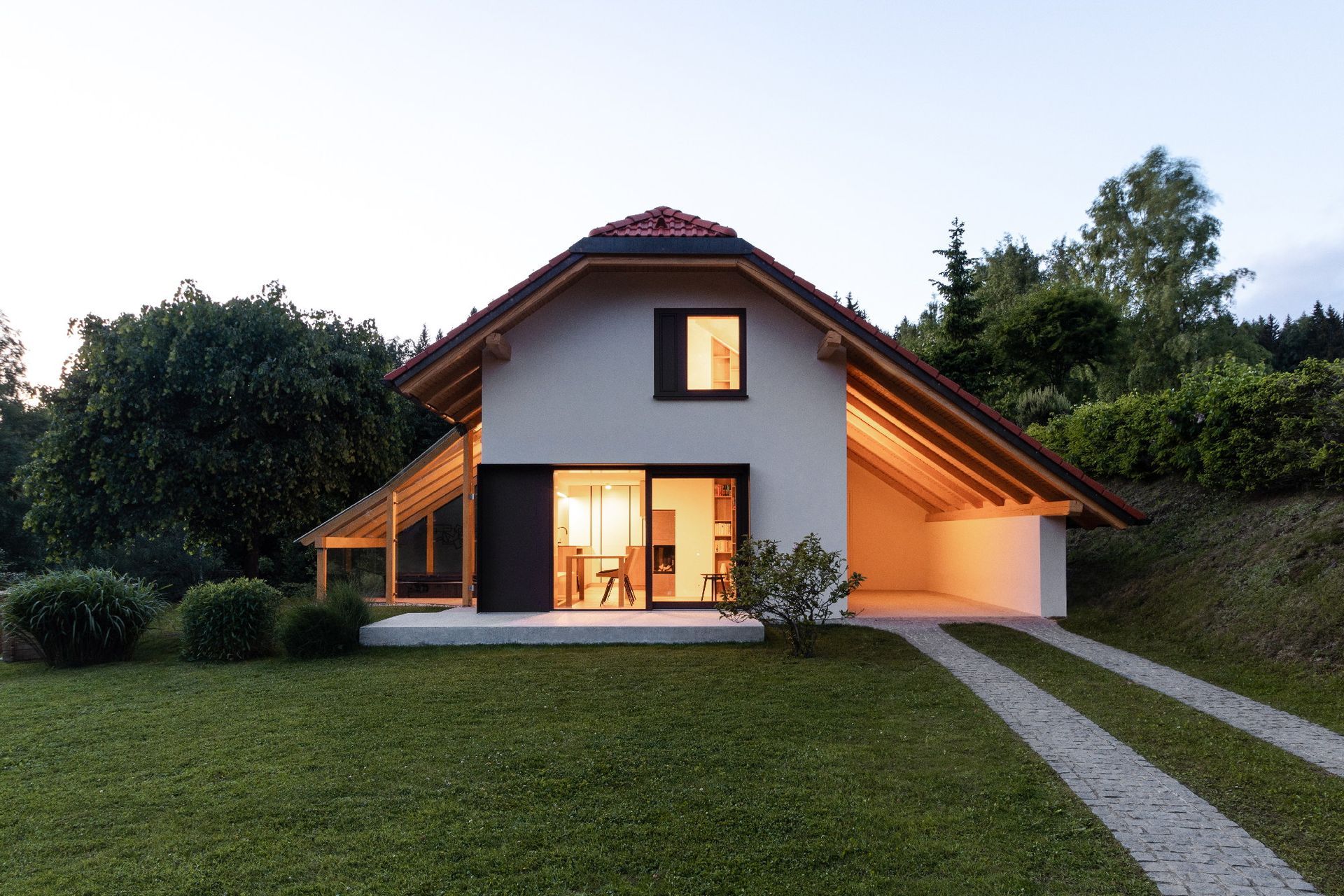 Casa moderna al crepuscolo con luci calde, circondata da erba e alberi, con un sentiero di pietra che conduce all'ingresso.
